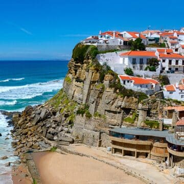 Vue sur une côte portuguaise, avec des maisons aux murs blancs à flanc de falaise. Une plage en premier plan et la mer en contre-bas.