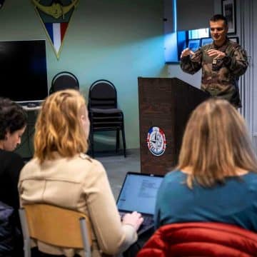 Estonian security officer standing at a desk, giving a conference to researchers sitting in a room.