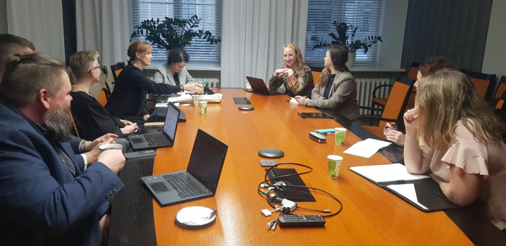 Eight people sitting around a meeting desk with computers and notepads on it.